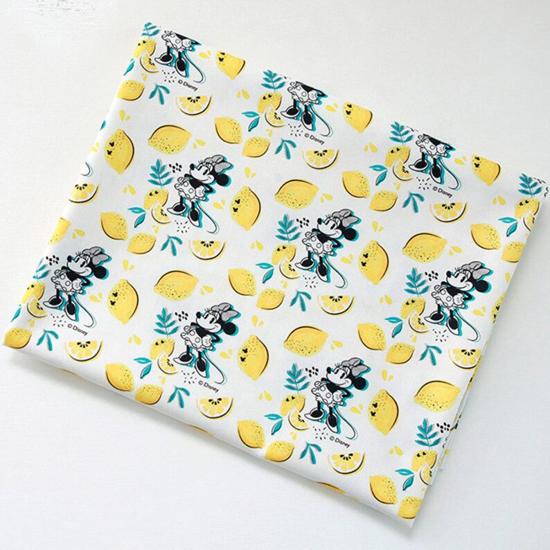 Puede incluir: Tela blanca con un patr&oacute;n repetido de Minnie Mouse y limones. Minnie Mouse lleva un vestido blanco y negro con un lazo rojo. Los limones son amarillos con hojas verdes. La tela tiene un fondo blanco con una textura sutil.