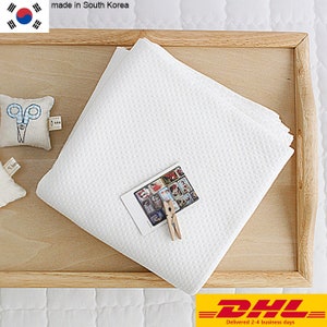 Peut inclure: Tissu blanc en nid d'abeille sur un plateau en bois. Le tissu est plié en trois et comporte une petite photo et une pince à linge sur le dessus. Le texte "made in South Korea" est visible dans le coin supérieur gauche de l'image.