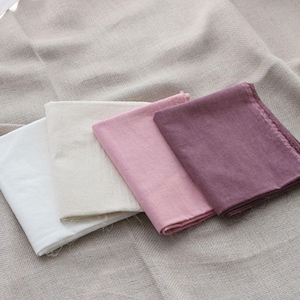 Peut inclure: Quatre carrés de tissu pliés disposés en rangée, présentant un dégradé de couleurs allant du blanc au crème, rose clair et mauve foncé. Chaque pièce de textile possède un tissage visible et des bords bruts, formant une composition simple.