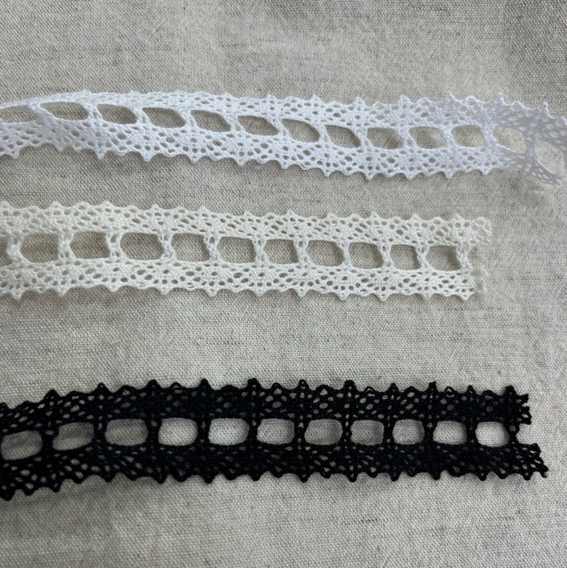 May include: Three pieces of lace trim, one white, one off-white, and one black. Each piece has a repeating pattern of diamond-shaped holes.