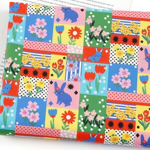 Puede incluir: Una tela con un colorido patrón de patchwork de bloques rectangulares. El diseño incluye conejos azules, tulipanes rojos, flores rosas y flores amarillas sobre fondos de color verde, amarillo, azul, rosa y lunares blancos y negros.