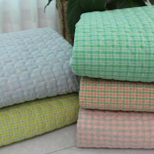 Może przedstawiać: Stos kolorowych, pikowanych koców. Koce są w odcieniach niebieskiego, zielonego, żółtego i różowego. Niektóre mają wzór w kratę, inne wzór w kształcie serca. Koce wydają się miękkie i wygodne.