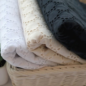 May include: Three rolls of white, beige, and black fabric with a delicate eyelet pattern. The fabric is folded and displayed in a wicker basket.