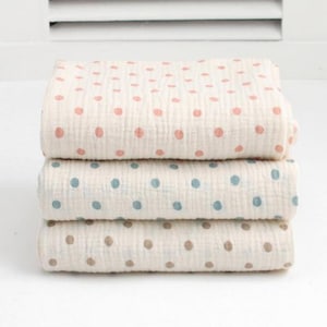 May include: A stack of three folded muslin blankets, each featuring a cream-colored base with a repeating polka dot pattern. The top blanket has pink dots, the middle blanket has blue dots, and the bottom blanket has brown dots.