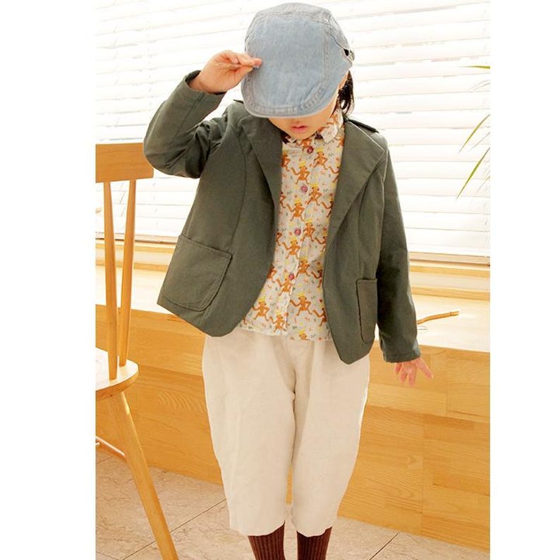 May include: A light blue denim flat cap, a dark olive green blazer with two front pockets, a white button-down shirt featuring a repeating orange monkey pattern, and plain white trousers. The outfit is worn as a complete set of clothing.