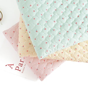 Peut inclure: Trois carrés de tissu matelassés de couleurs différentes : vert clair, jaune et rose. Chaque carré présente un petit motif de cerises rouges et un motif de grille blanche.