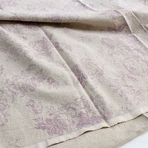 K&ouml;nnte beinhalten: Ein beigefarbener Stoff mit einem sich wiederholenden Blumenmuster in einem ged&auml;mpften Violettton. Das Textil weist eine sichtbare Webstruktur mit weichen Falten und einer rohen, unvers&auml;uberten Kante am unteren Rand auf.