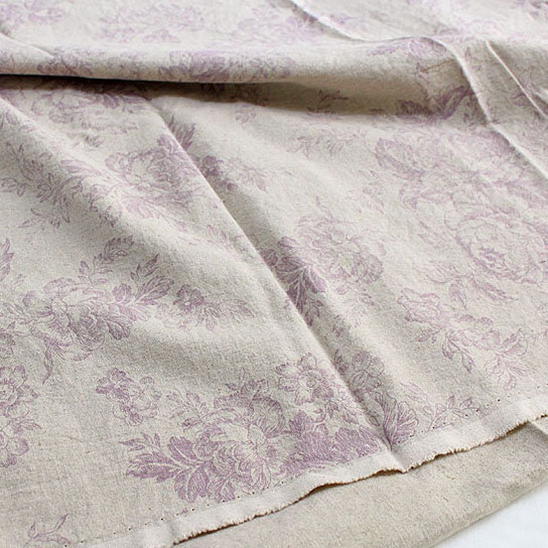 K&ouml;nnte beinhalten: Ein beigefarbener Stoff mit einem sich wiederholenden Blumenmuster in einem ged&auml;mpften Violettton. Das Textil weist eine sichtbare Webstruktur mit weichen Falten und einer rohen, unvers&auml;uberten Kante am unteren Rand auf.