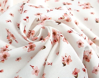 May include: A white fabric featuring a repeating pattern of small, light pink cherry blossom flowers with brown centers and stems. The textile has a visible woven texture and is gathered in the center to show the drape and folds of the material.