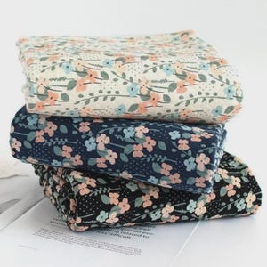 May include: Three floral patterned fabrics stacked on top of each other. The top fabric is cream with peach and blue flowers. The middle fabric is navy with peach and blue flowers. The bottom fabric is black with peach and blue flowers.
