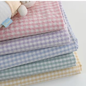 May include: A stack of five folded fabric squares featuring a gingham check pattern in pastel pink, lavender, light blue, mint green, and soft yellow. Each square has a textured, crinkled surface and a small blue tag attached to the top layer.