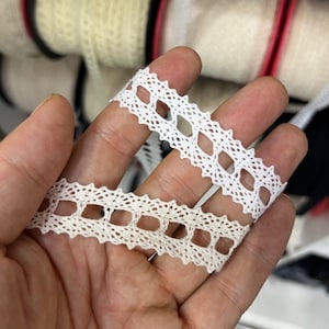 May include: Two white lace trims with a delicate square pattern. The lace is shown on a hand.