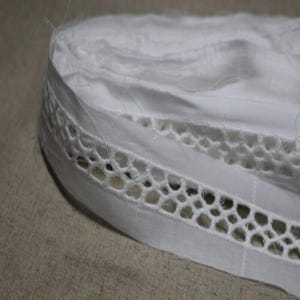 May include: Close-up of white fabric with a decorative openwork trim. The trim has a repeating pattern of interconnected, rounded shapes. The fabric appears layered, with visible stitching and a slightly textured surface. The overall aesthetic is delicate and possibly vintage.
