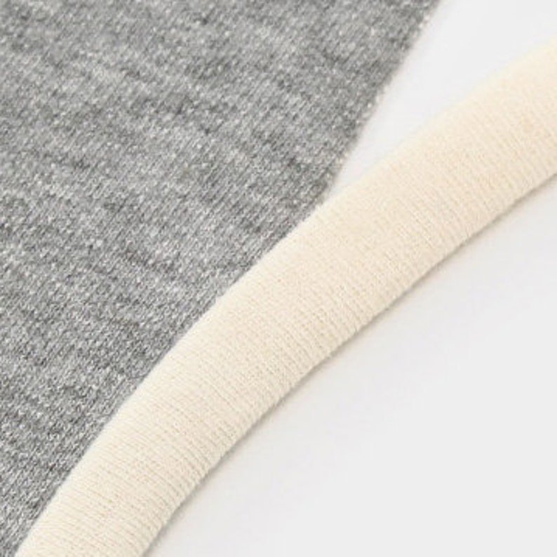 Pu&ograve; includere: Una vista ravvicinata di un tessuto grigio m&eacute;lange con un motivo a maglia fine e testurizzato, caratterizzato da un bordo o una rifinitura a coste spessa di colore crema lungo il bordo curvo. Il materiale appare morbido e flessibile, mostrando un netto contrasto tra il tessuto grigio e il bordo chiaro.