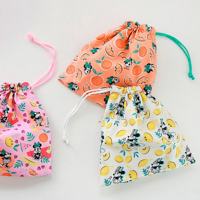 Puede incluir: Tres bolsas de cord&oacute;n con diferentes patrones de frutas. La primera bolsa es rosa con fondo blanco y presenta un patr&oacute;n de frutas rosas y amarillas, hojas verdes y personajes de Mickey Mouse. La segunda bolsa es naranja con fondo blanco y presenta un patr&oacute;n de rodajas de naranja y personajes de Mickey Mouse. La tercera bolsa es blanca con fondo amarillo y presenta un patr&oacute;n de limones y personajes de Mickey Mouse.