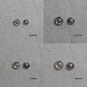 May include: Four sets of silver metal snap fasteners in different sizes: 12mm, 14mm, 17mm, and 19mm. The snaps are arranged in rows on a light grey fabric background.