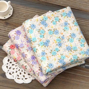 May include: Three folded floral fabric pieces with a white lace doily and a teacup on a wooden surface. The fabrics feature a repeating pattern of blue, purple, and pink roses on a cream background. The teacup is white.