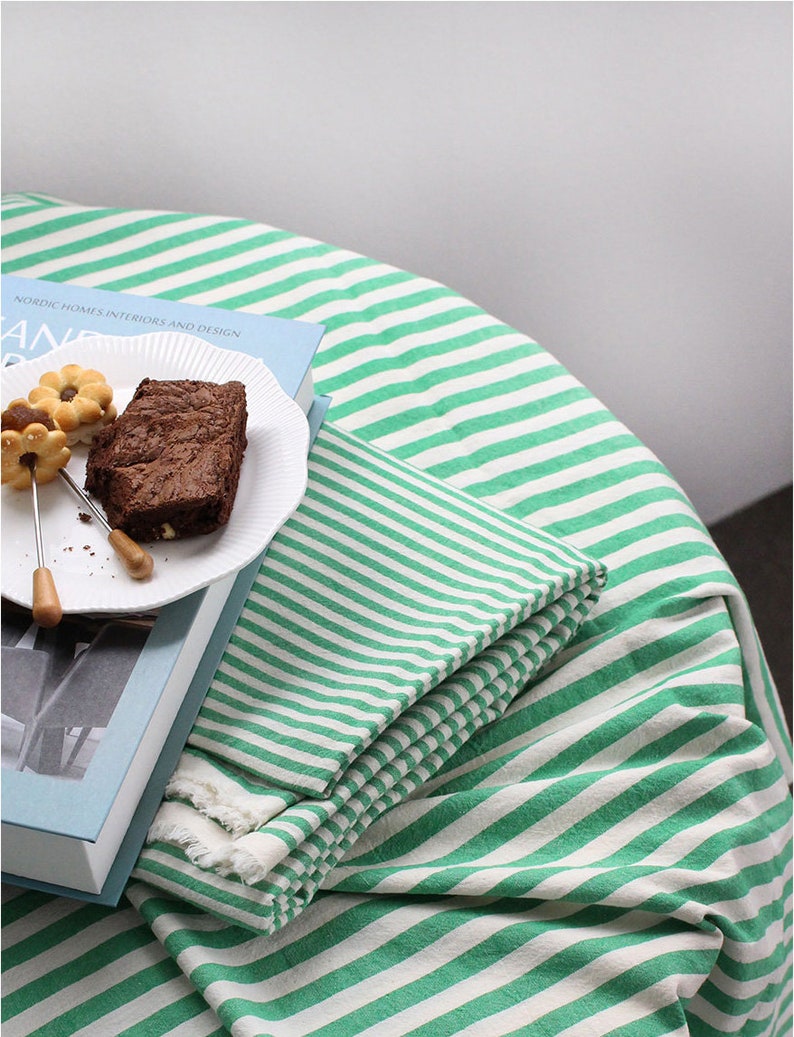 K&ouml;nnte beinhalten: Eine gr&uuml;n-wei&szlig; gestreifte Tischdecke mit einem Buch und einem Teller Brownies darauf. Das Buch hat den Text "Nordic Homes Interiors and Design" auf dem Cover.