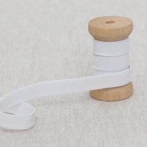 May include: White elastic ribbon spooled on a wooden bobbin. The ribbon is unwound, showing its flat, woven texture. The bobbin is made of light-colored wood, and the background is a neutral fabric.