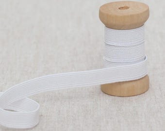 May include: White elastic ribbon spooled on a wooden bobbin. The ribbon is unwound, showing its flat, woven texture. The bobbin is made of light-colored wood, and the background is a neutral fabric.