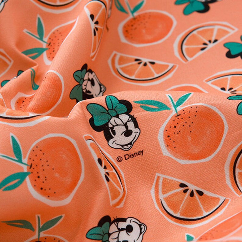 Puede incluir: Tela de color melocot&oacute;n con un patr&oacute;n repetido de rodajas de naranja e im&aacute;genes de dibujos animados de Minnie Mouse con un lazo verde. La tela tiene el texto "&copy; Disney" impreso.