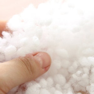 May include: Close-up of a hand holding a mass of fluffy, white polyester fiberfill. The soft, cloud-like material is tightly packed and appears to be used for stuffing pillows, cushions, or other craft projects. The background is a soft, neutral color.