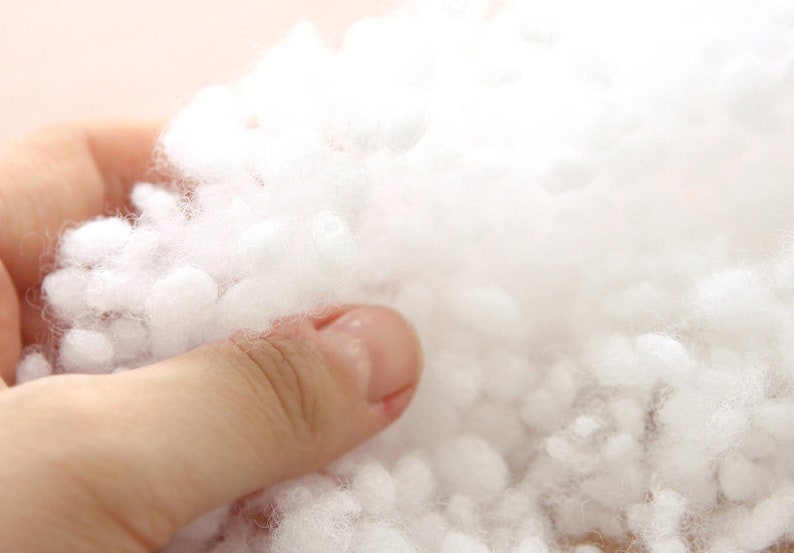 May include: Close-up of a hand holding a mass of fluffy, white polyester fiberfill. The soft, cloud-like material is tightly packed and appears to be used for stuffing pillows, cushions, or other craft projects. The background is a soft, neutral color.