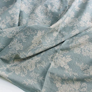 K&ouml;nnte beinhalten: Ein hellblauer Stoff mit einem sich wiederholenden wei&szlig;en Blumenmuster aus detaillierten Bl&uuml;ten und Bl&auml;ttern. Das Textil hat eine weiche, matte Textur mit sichtbaren Falten und Drapierungen, die Tiefe auf der Materialoberfl&auml;che erzeugen.