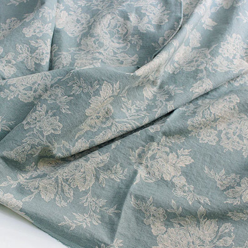 K&ouml;nnte beinhalten: Ein hellblauer Stoff mit einem sich wiederholenden wei&szlig;en Blumenmuster aus detaillierten Bl&uuml;ten und Bl&auml;ttern. Das Textil hat eine weiche, matte Textur mit sichtbaren Falten und Drapierungen, die Tiefe auf der Materialoberfl&auml;che erzeugen.