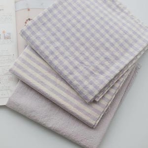 May include: Three folded pieces of fabric stacked together. The top piece features a light purple and white gingham check pattern. The middle piece has a light purple and white striped pattern. The bottom piece is a solid light purple fabric.