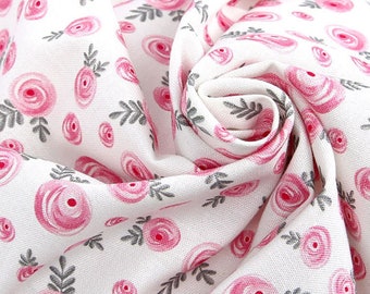 May include: A white fabric featuring a repeating pattern of pink circular rose motifs and small grey leaf sprigs. The textile is gathered into a soft, swirling fold, highlighting the texture of the woven material and the floral print design.