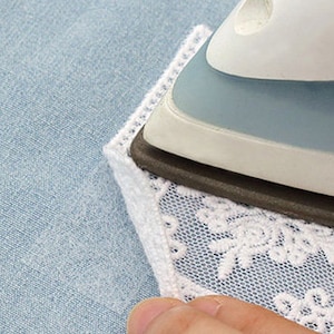 May include: A close-up of a white lace patch being ironed onto a light blue fabric with a blue and white iron. The lace has a delicate floral pattern and is being pressed flat.
