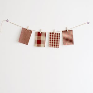 May include: Four fabric swatches in shades of red and beige are clipped to a string with wooden clothespins. The swatches display solid, plaid, and gingham patterns. The string is suspended against a white background, creating a simple, rustic display.