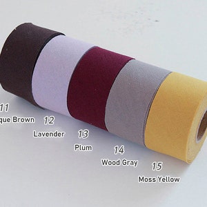 May include: A horizontal roll of fabric tape divided into five distinct colour sections labelled with numbers and names: 11 Antique Brown, 12 Lavender, 13 Plum, 14 Wood Gray, and 15 Moss Yellow. The tape has a smooth, matte texture and a consistent width.