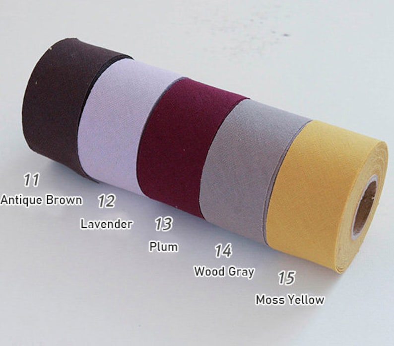May include: A horizontal roll of fabric tape divided into five distinct colour sections labelled with numbers and names: 11 Antique Brown, 12 Lavender, 13 Plum, 14 Wood Gray, and 15 Moss Yellow. The tape has a smooth, matte texture and a consistent width.
