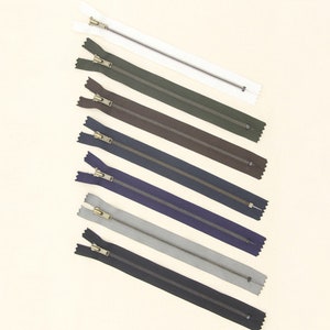 May include: Assortment of eight zipper closures in various colors, including white, green, brown, navy, gray, and black. The zippers have brass pulls.