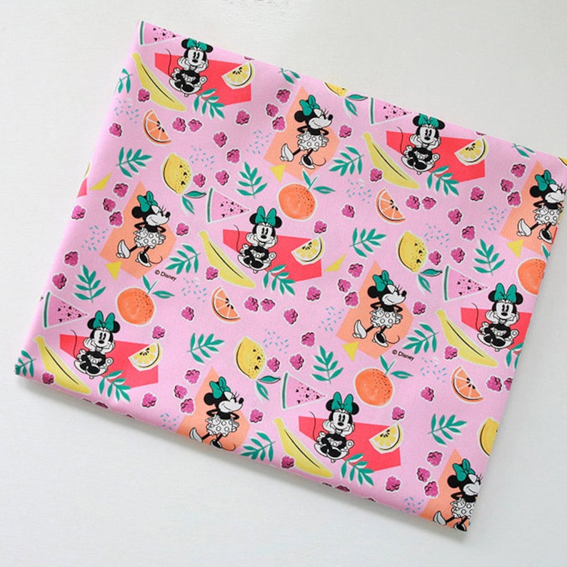 Puede incluir: Tela rosa con un patr&oacute;n repetido de Minnie Mouse, pl&aacute;tanos, limones, naranjas y frambuesas. El fondo es rosa con un patr&oacute;n de lunares blancos. La tela est&aacute; impresa con el texto "&copy; Disney".
