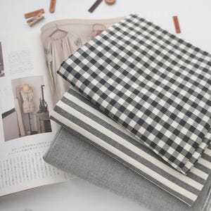 Peut inclure: Trois échantillons de tissu superposés. Le premier est à carreaux noirs et blancs, le second à rayures grises et blanches, et le troisième est gris uni. Ils sont posés sur un magazine ouvert.