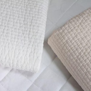 May include: Two folded, quilted blankets in the image. One is bright white, and the other is a light beige color. The blankets have a textured, grid-like pattern. The background is a white, quilted surface.