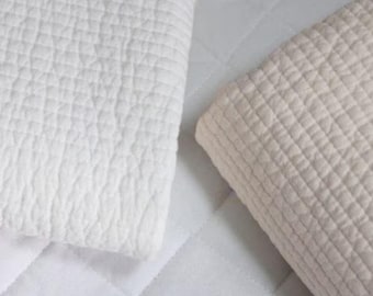 Puede incluir: Dos mantas acolchadas y plegadas. Una es blanca, la otra de color beige claro. Ambas tienen un patrón texturizado en forma de rejilla. El fondo es una superficie acolchada blanca. La imagen está bien iluminada.