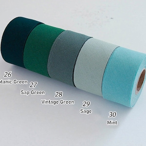May include: Five rolls of fabric ribbon arranged in a row, featuring a gradient of green shades labelled 26 Botanic Green, 27 Sap Green, 28 Vintage Green, 29 Sage, and 30 Mint. Each roll has a textured surface and a central cardboard core.