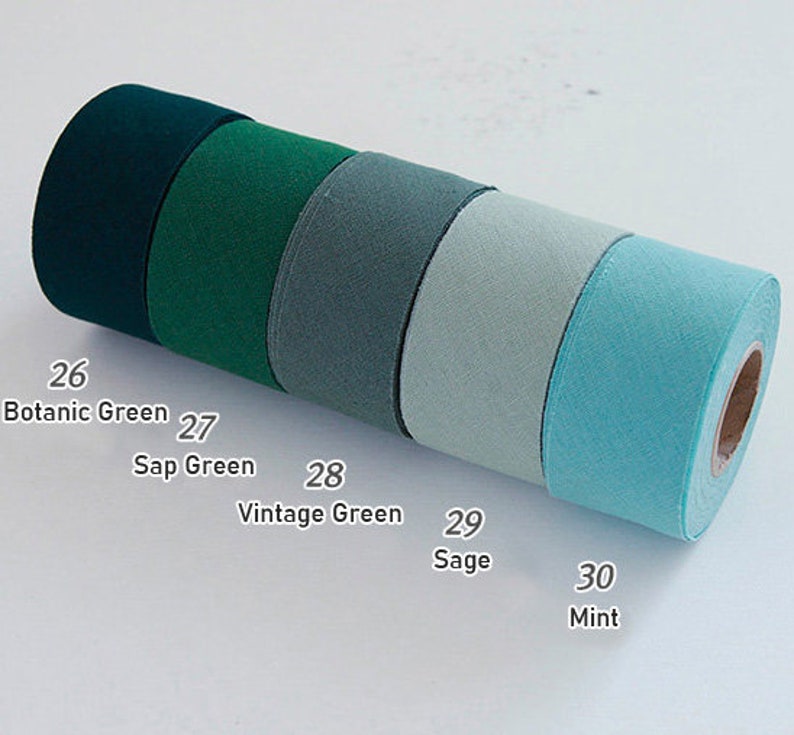 May include: Five rolls of fabric ribbon arranged in a row, featuring a gradient of green shades labelled 26 Botanic Green, 27 Sap Green, 28 Vintage Green, 29 Sage, and 30 Mint. Each roll has a textured surface and a central cardboard core.
