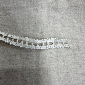 May include: White lace trim with a delicate, openwork design. The lace is on a beige fabric background.