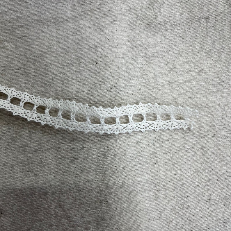 May include: White lace trim with a delicate, openwork design. The lace is on a beige fabric background.