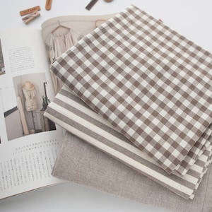 May include: A stack of three folded fabric pieces featuring a brown and white gingham check pattern, a brown and white striped pattern, and a solid light brown textile. The fabrics are layered on top of an open magazine with Japanese text.