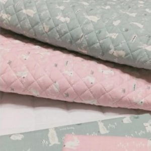 May include: Close-up of quilted fabric in shades of pink and green. The fabric features a repeating pattern of white bunnies and small shapes. The quilting creates a diamond pattern across the surface. The fabric appears soft and suitable for baby items.