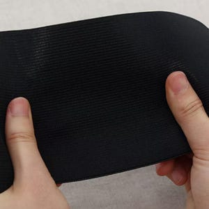 May include: Close-up of a black elastic band with a textured surface. The band is held between two hands, showcasing its flexibility and material. The fabric appears to be tightly woven, suggesting durability and stretch.