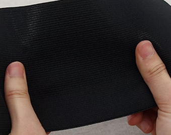 May include: Close-up of a black elastic band with a textured surface. The band is held between two hands, showcasing its flexibility and material. The fabric appears to be tightly woven, suggesting durability and stretch.