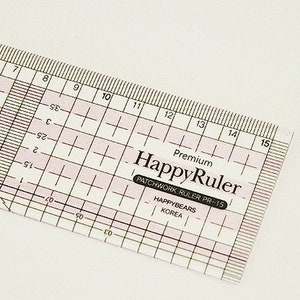 Puede incluir: Una regla HappyRuler de pl&aacute;stico transparente con l&iacute;neas de cuadr&iacute;cula rosas y medidas en cent&iacute;metros. La regla tiene el texto "HappyRuler" y "PATCHWORK RULER PR-15" impreso. La regla mide unos 38 cm.