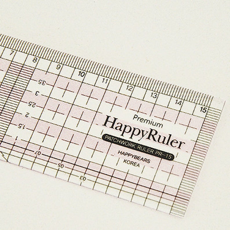 Puede incluir: Una regla HappyRuler de pl&aacute;stico transparente con l&iacute;neas de cuadr&iacute;cula rosas y medidas en cent&iacute;metros. La regla tiene el texto "HappyRuler" y "PATCHWORK RULER PR-15" impreso. La regla mide unos 38 cm.